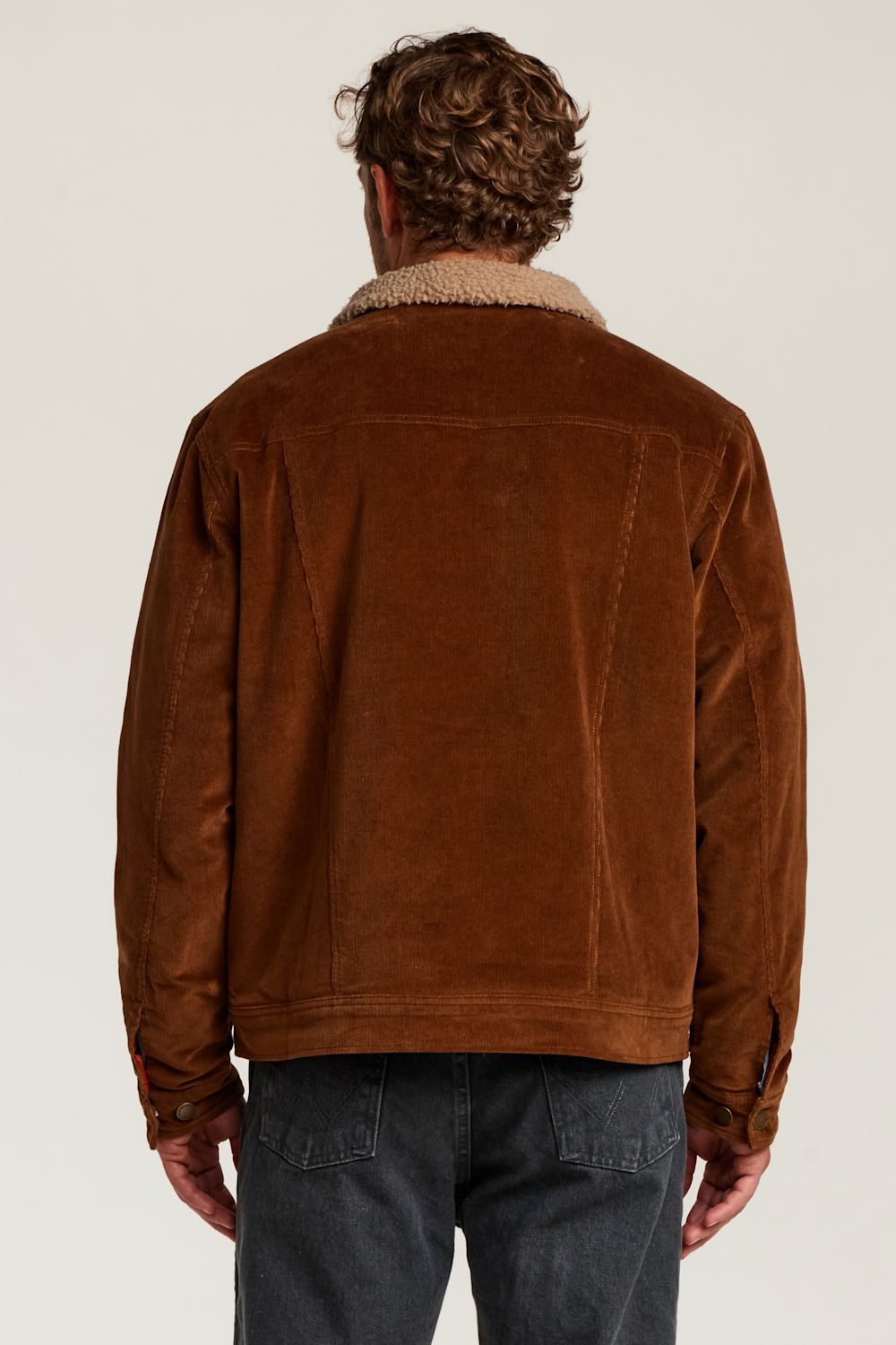 A man viewed from behind wearing a brown corduroy jacket with a beige sherpa collar and dark gray jeans, set against a plain light gray studio background.