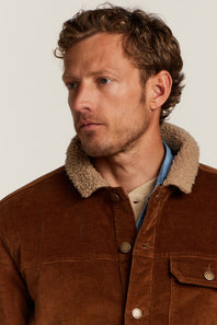 A close-up of a man wearing a brown corduroy jacket with a beige sherpa collar, layered over a light blue shirt and beige t-shirt, against a plain off-white background.