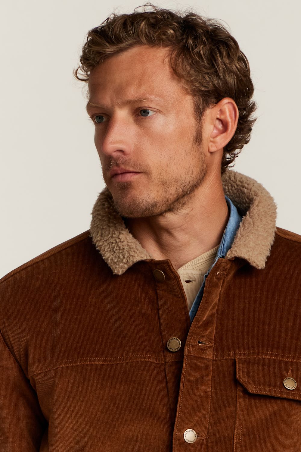 A close-up of a man wearing a brown corduroy jacket with a beige sherpa collar, layered over a light blue shirt and beige t-shirt, against a plain off-white background.