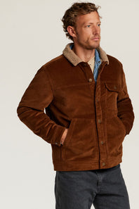 A man wearing a brown corduroy jacket with a beige sherpa collar, layered over a blue shirt and dark jeans, photographed against a light gray studio background.