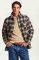 A man wearing a brown, black, and beige plaid flannel shirt jacket over a beige henley shirt, paired with blue jeans and a brown patterned belt, against a light off-white background.