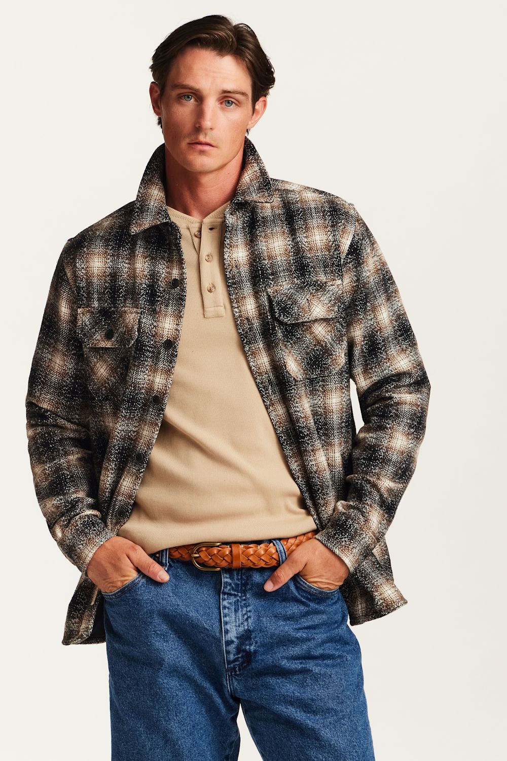 A man wearing a brown, black, and beige plaid flannel shirt jacket over a beige henley shirt, paired with blue jeans and a brown patterned belt, against a light off-white background.