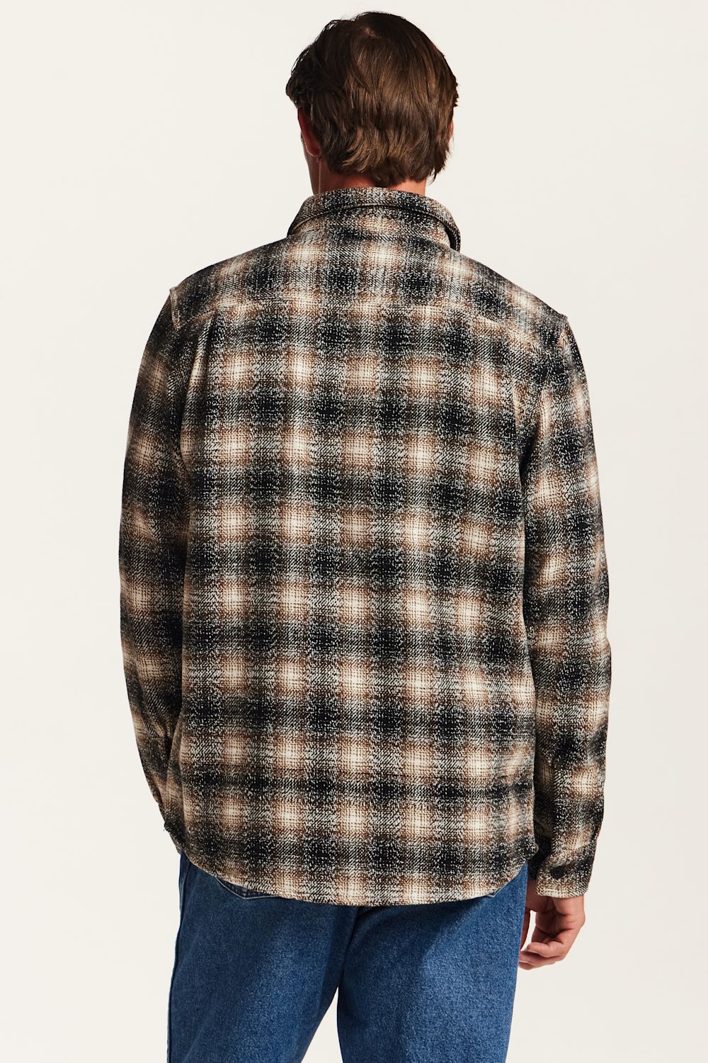 A man viewed from behind wearing a long-sleeve plaid button-up shirt in black, brown, and beige, paired with blue jeans, against a plain off-white background.