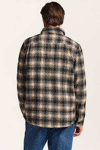 A man viewed from behind wearing a long-sleeve plaid button-up shirt in black, brown, and beige, paired with blue jeans, against a plain off-white background.