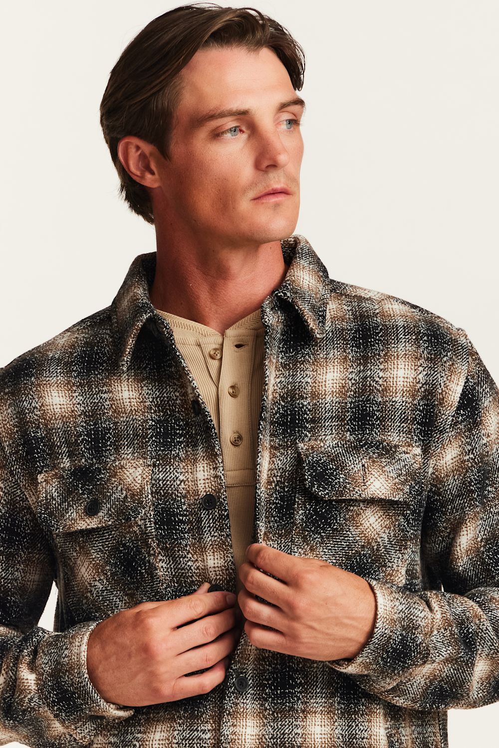 A man wearing a brown, black, and beige plaid flannel overshirt over a tan henley shirt, photographed against a light off-white background.