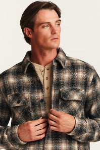 A man wearing a brown, black, and beige plaid flannel overshirt over a tan henley shirt, photographed against a light off-white background.