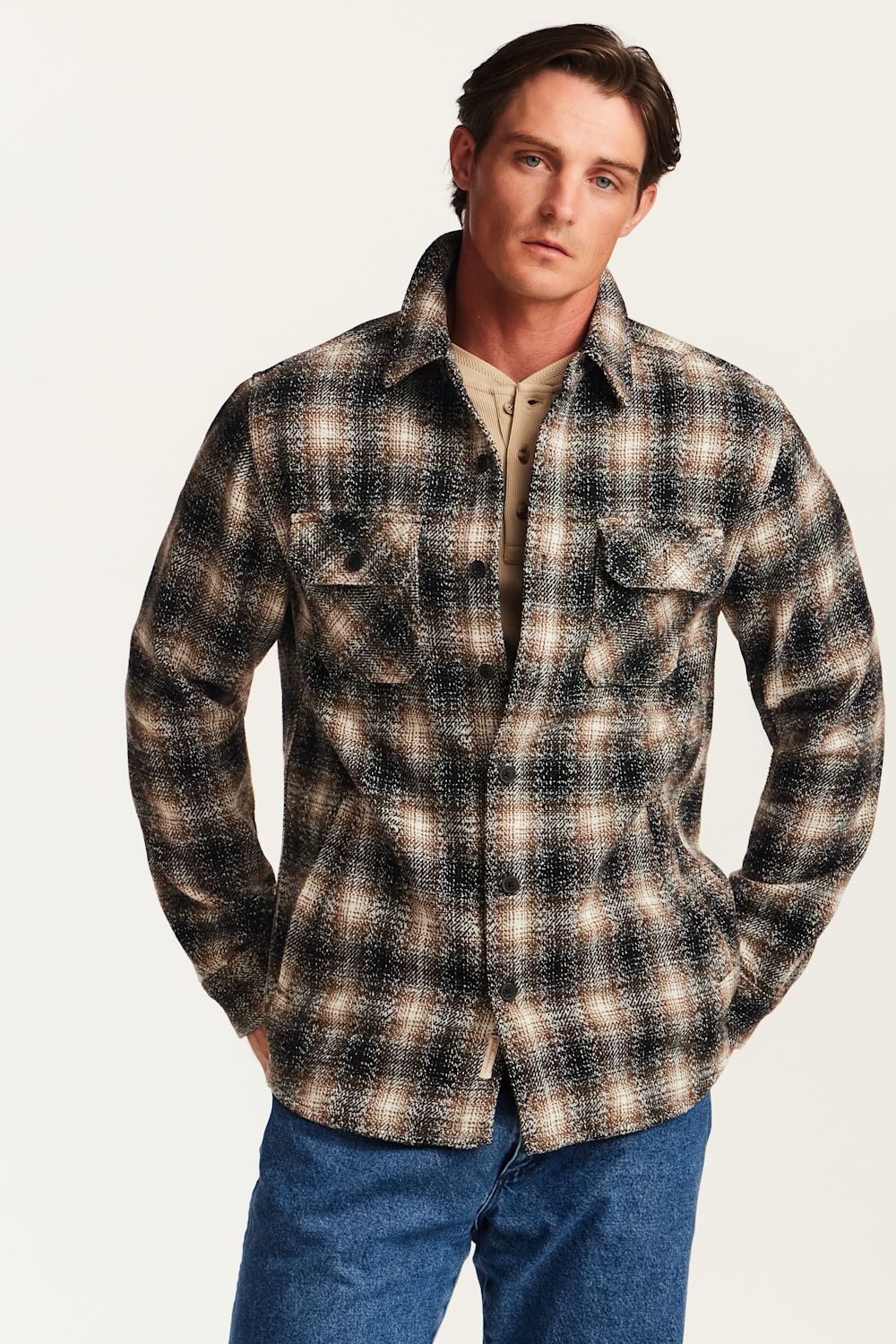 A man wearing a long-sleeve plaid button-up shirt in brown, beige, and black tones, paired with blue jeans, against a plain light background.