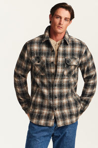 A man wearing a long-sleeve plaid button-up shirt in brown, beige, and black tones, paired with blue jeans, against a plain light background.