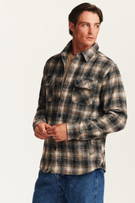A man wearing a long-sleeve button-up plaid flannel shirt in black, brown, and beige, paired with blue jeans, photographed against a clean off-white studio background.