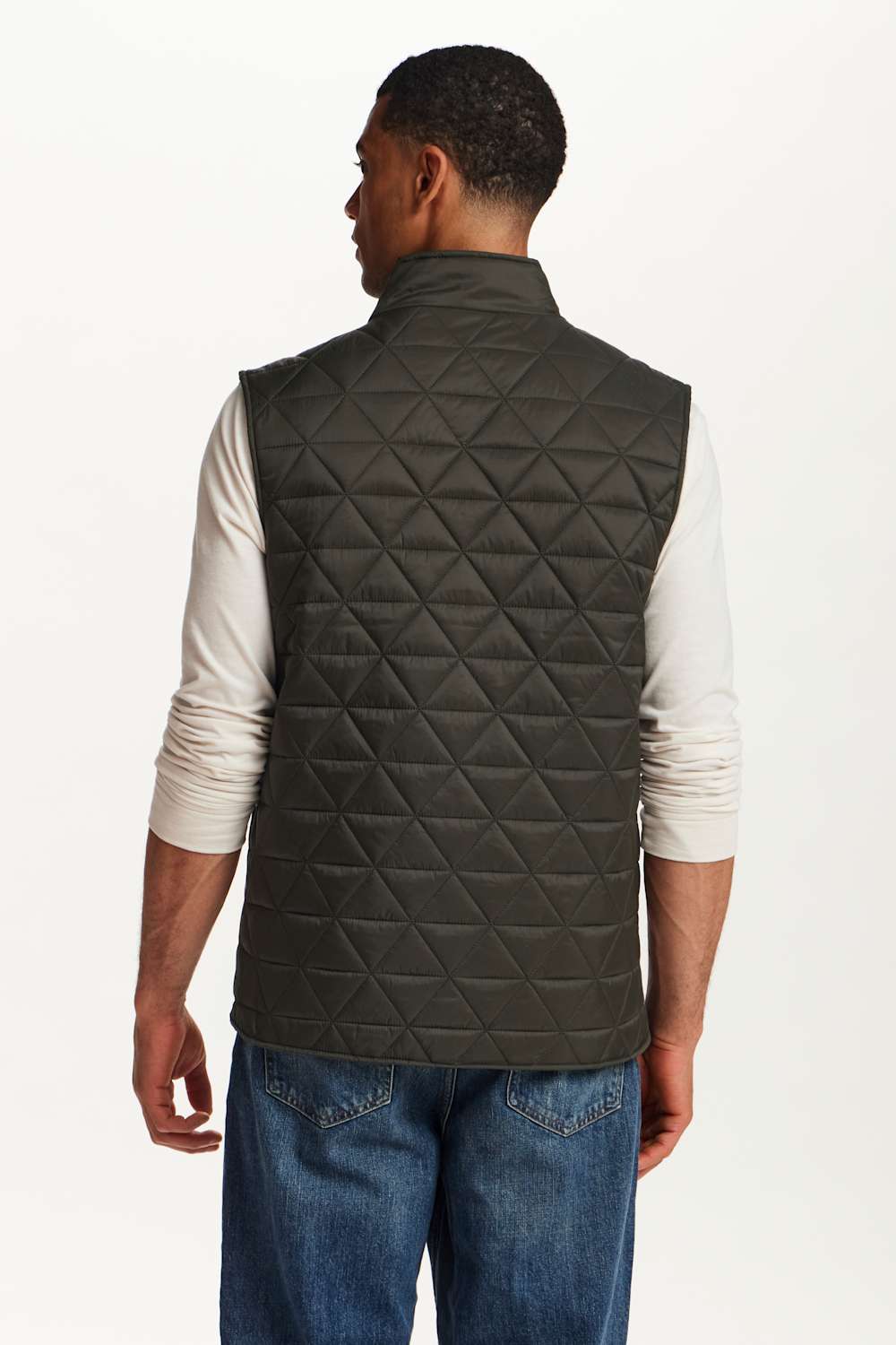 A man viewed from the back wearing a dark olive green diamond-quilted puffer vest over a white long-sleeve shirt and blue jeans, against a plain white studio background.