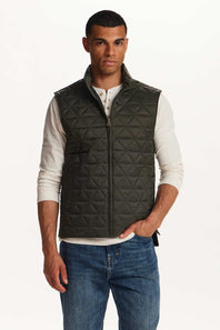 A man wearing a dark olive green diamond-quilted zip-up vest over a white henley shirt and blue jeans, standing against a plain white studio background.