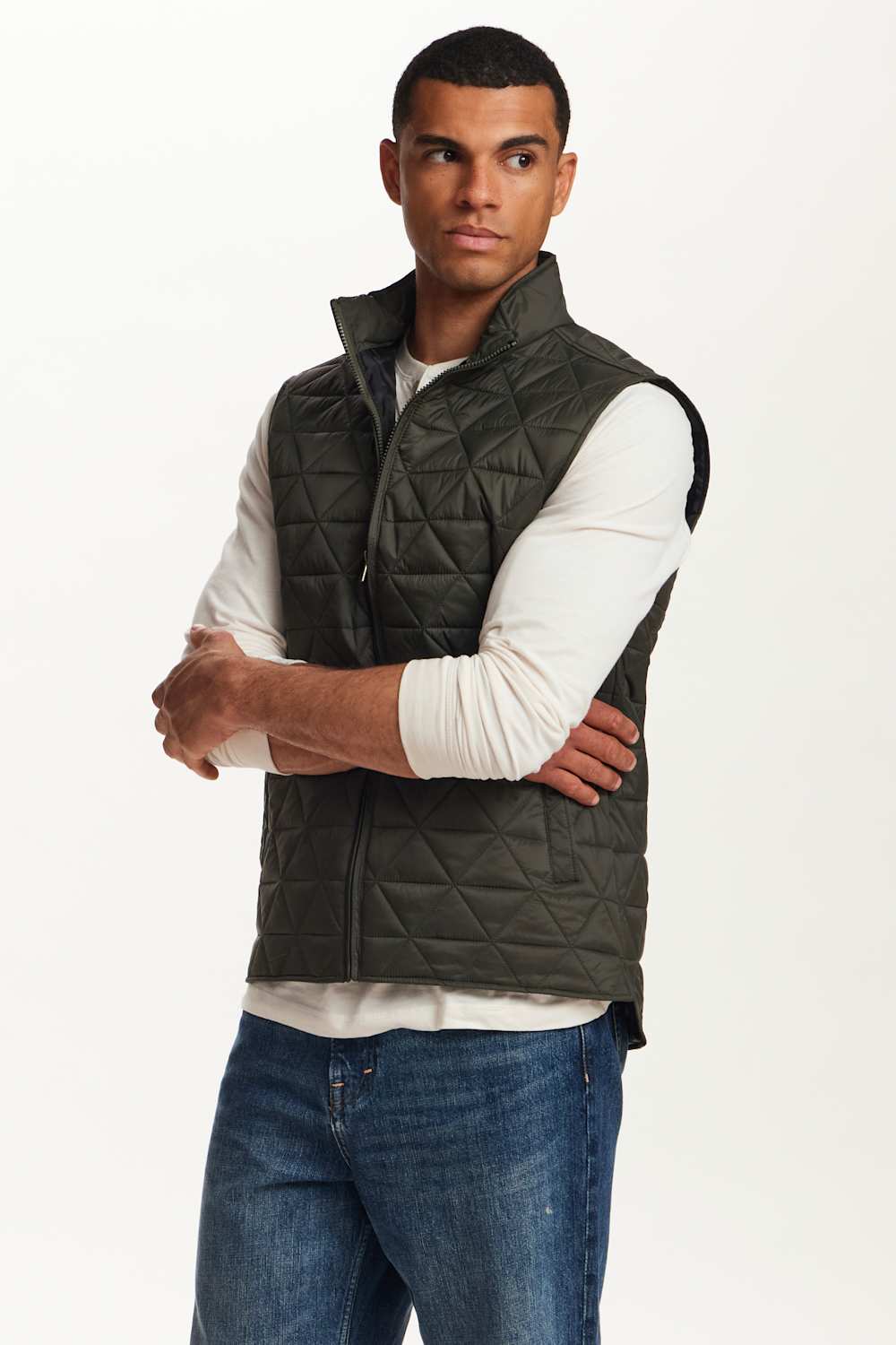 A man wearing an olive green diamond-quilted zip-up puffer vest over a white long-sleeve shirt and blue jeans, standing against a plain white studio background.