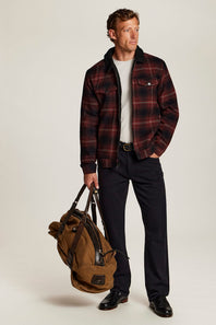 A man stands against a light beige studio background wearing a dark red and black plaid overshirt over a white t-shirt, dark trousers, and black shoes, while carrying a brown duffel bag.