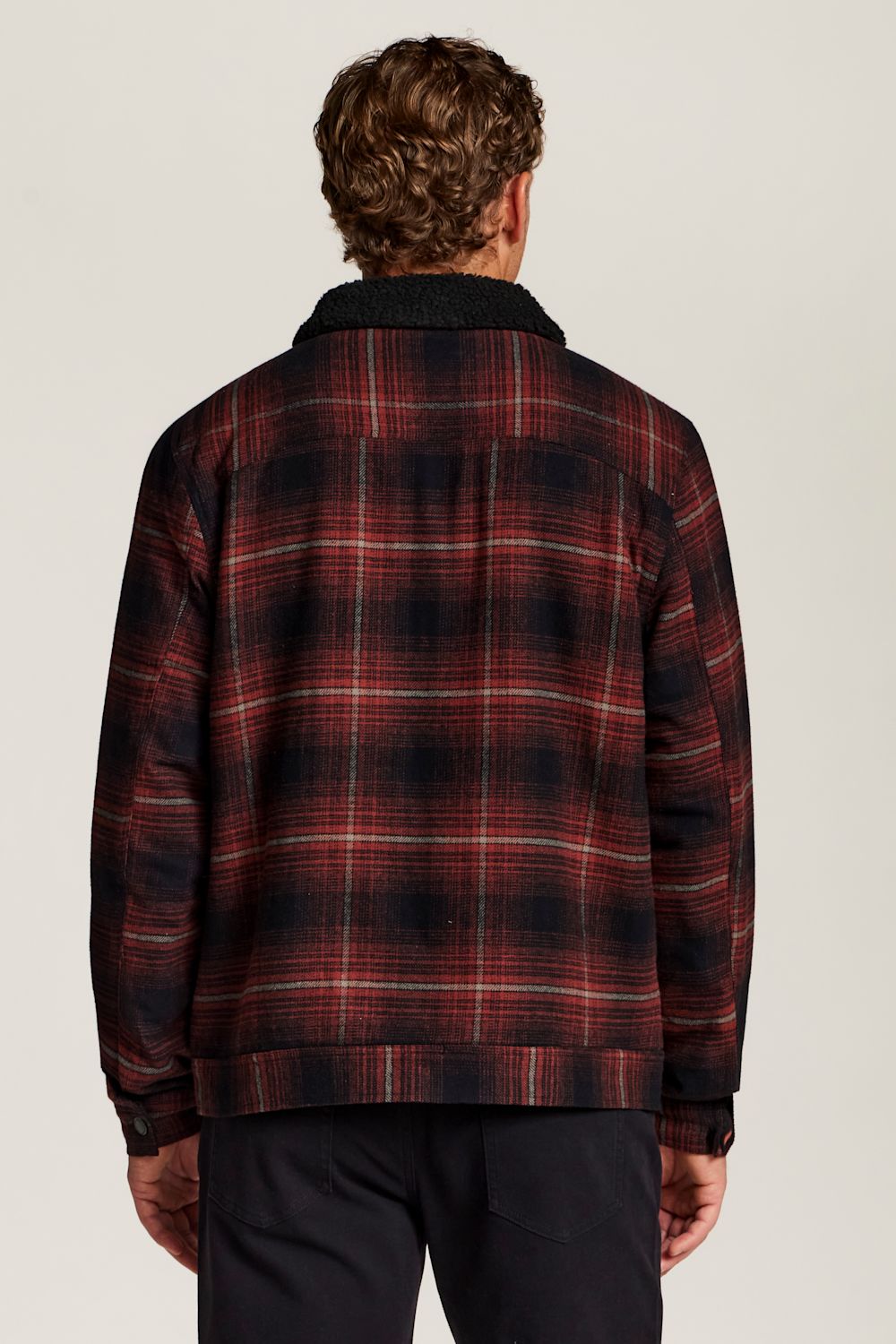 A man viewed from behind wearing a red and black plaid flannel jacket with a dark corduroy collar, paired with dark trousers, against a light gray studio background.