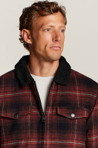 A man wearing a red and black plaid zip-up jacket with a black sherpa collar, layered over a white t-shirt, against a light gray background.