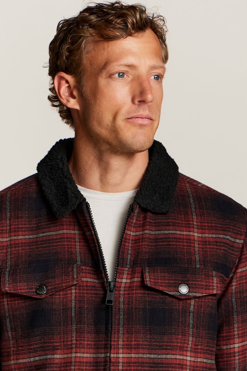 A man wearing a red and black plaid zip-up jacket with a black sherpa collar, layered over a white t-shirt, against a light gray background.