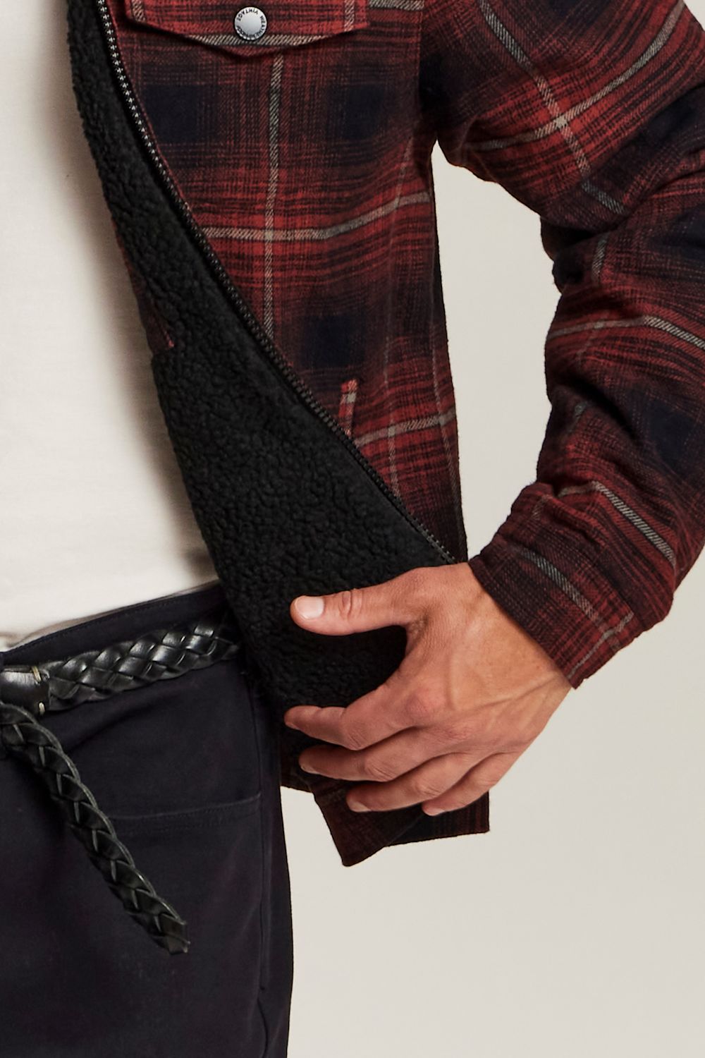 Close-up of a person wearing a red and black plaid overshirt with a black sherpa lining, paired with dark trousers and a black braided belt, against a light neutral background.