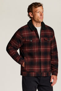 A man wearing a red and black plaid zip-up jacket with a dark sherpa collar, standing against a light gray studio background.