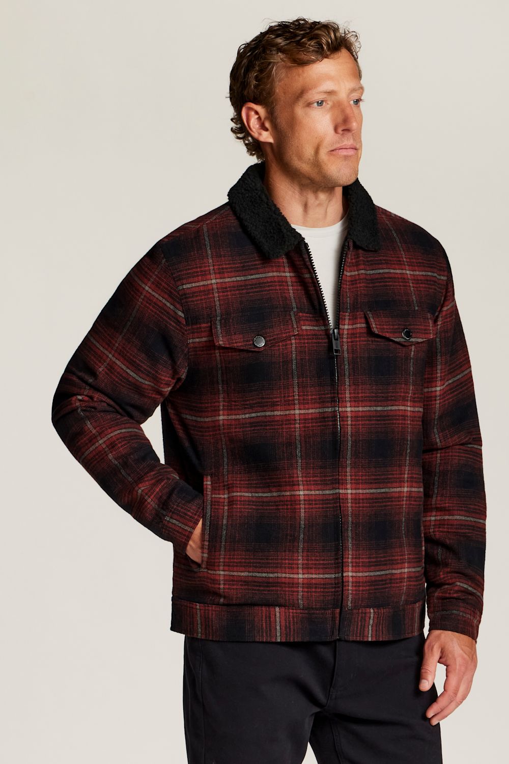A man wearing a red and black plaid zip-up jacket with a dark sherpa collar, standing against a light gray studio background.