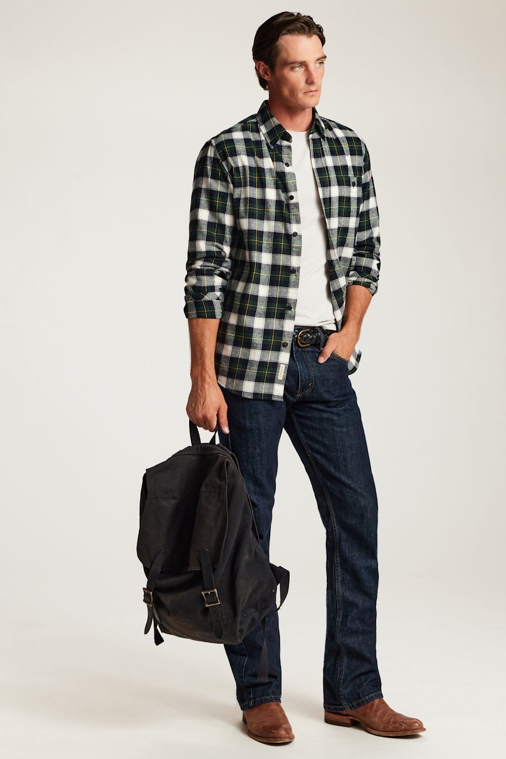 A man modeling a green and black plaid flannel shirt over a white t-shirt, paired with dark blue jeans and brown leather boots, while holding a black backpack against a light gray studio background.