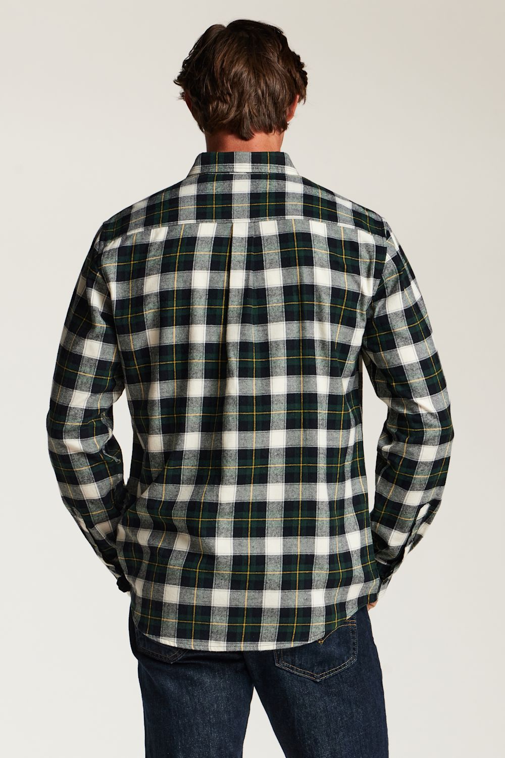 A man viewed from behind wearing a long-sleeve plaid flannel shirt in green, white, and yellow, paired with dark blue jeans, against a light gray studio background.