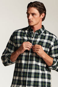 A man wearing a long-sleeve button-down plaid shirt in dark green, white, and yellow, adjusting the buttons on the front. The photo is taken against a plain light gray studio background.