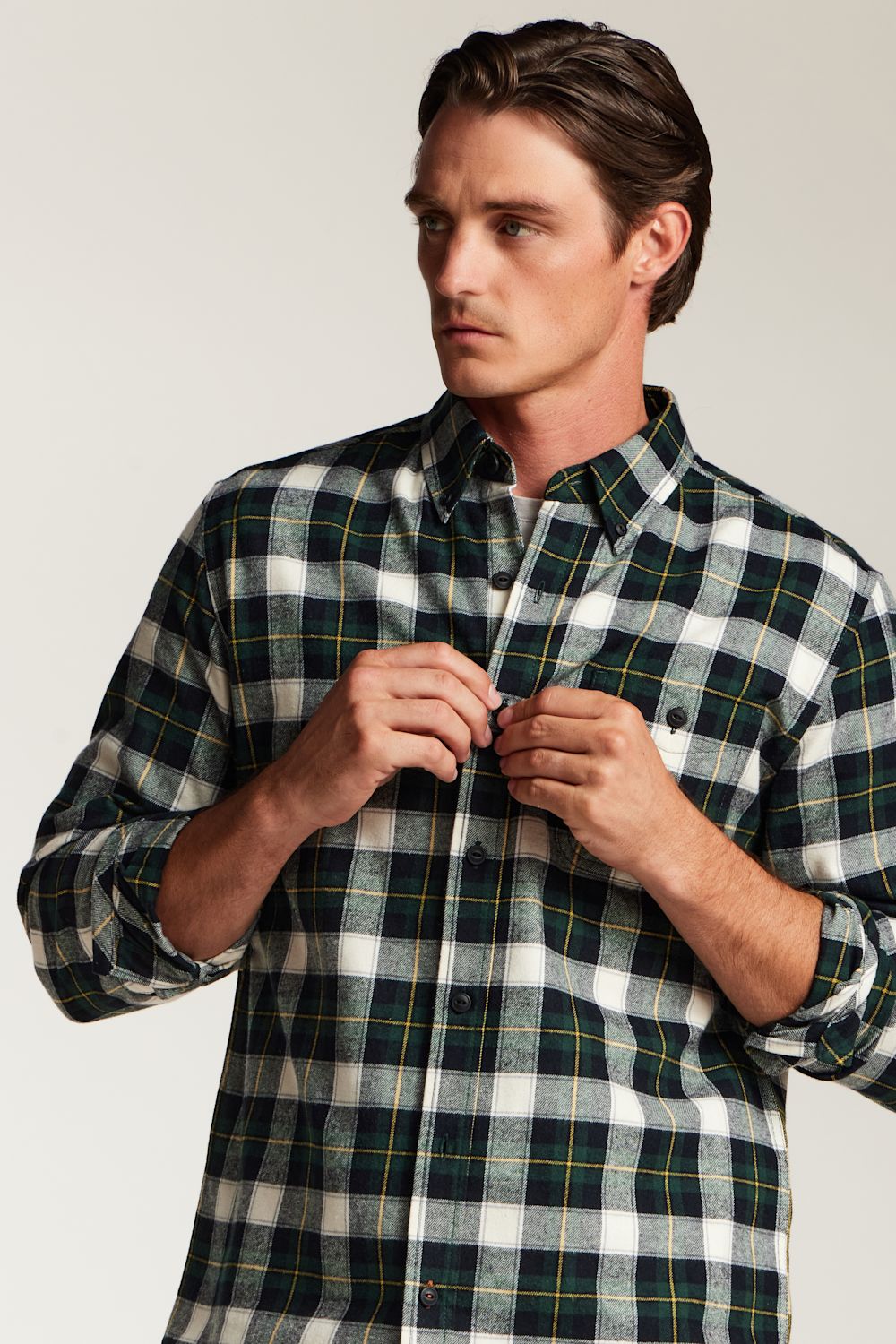 A man wearing a long-sleeve button-down plaid shirt in dark green, white, and yellow, adjusting the buttons on the front. The photo is taken against a plain light gray studio background.