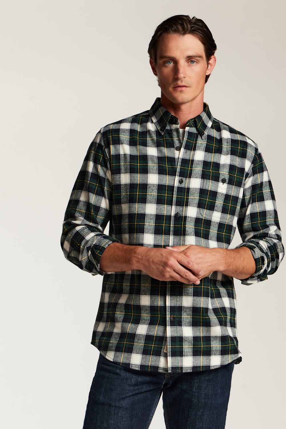 A man wearing a long-sleeve button-down plaid shirt in green, white, and yellow, paired with dark blue jeans, photographed against a light gray studio background.