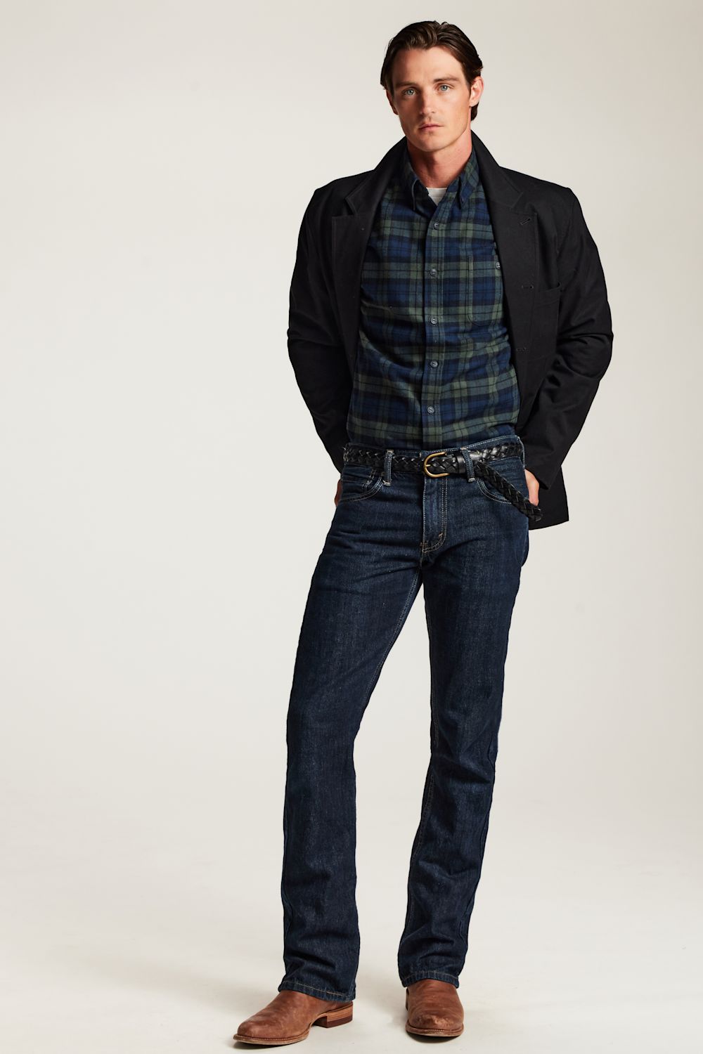 A man modeling a dark navy jacket, a blue and green plaid button-down shirt, dark wash jeans, and brown leather shoes against a plain light gray studio background.
