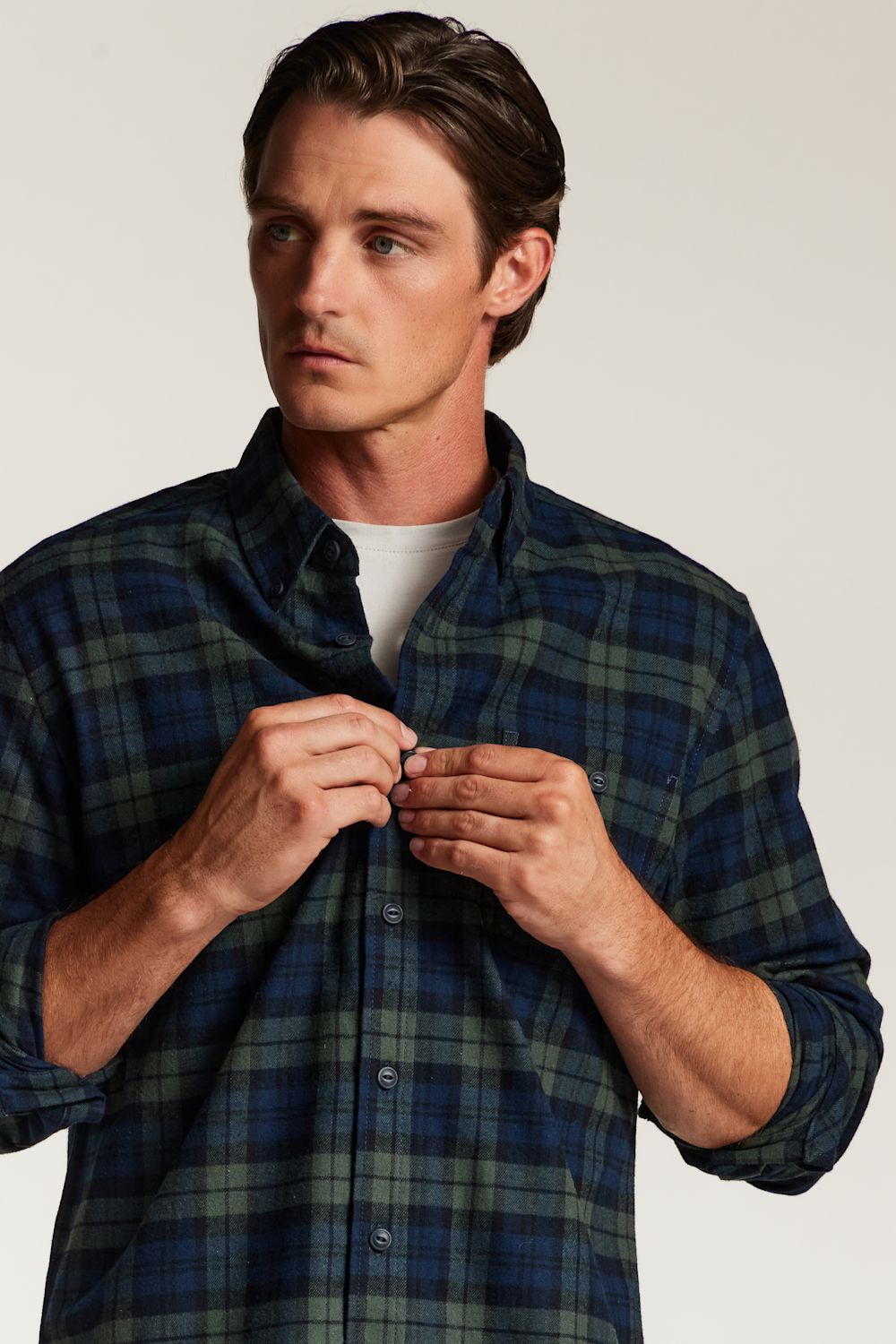A man wearing a dark blue and green plaid button-down shirt over a white t-shirt, adjusting the front button. The photo is taken against a plain light gray studio background.