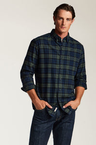 A man wearing a dark navy and green plaid button-down shirt with rolled-up sleeves, paired with dark blue jeans, standing against a light gray studio background.