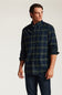 A man wearing a dark navy and green plaid button-down shirt with dark blue jeans, standing against a plain light gray studio background.