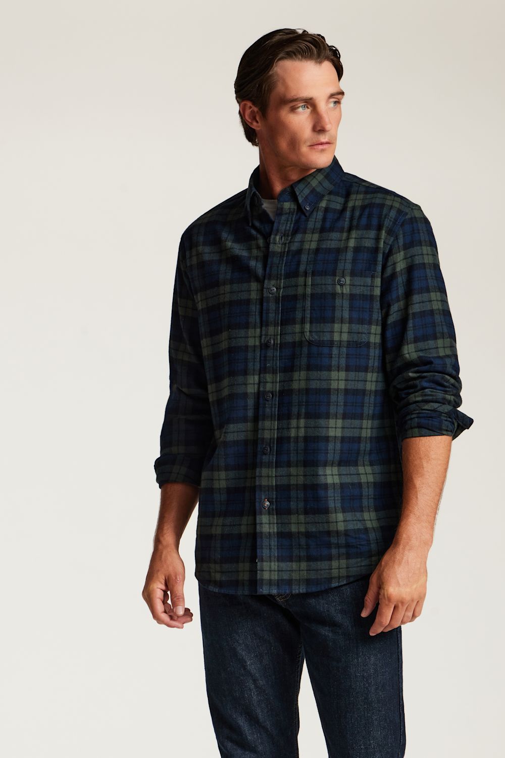 A man wearing a dark navy and green plaid button-down shirt with dark blue jeans, standing against a plain light gray studio background.