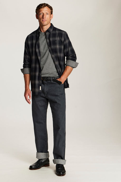 A man modeling a dark gray and black plaid overshirt over a gray t-shirt, paired with dark gray jeans and black shoes, against a plain light gray studio background.