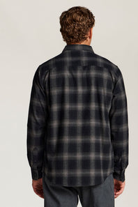 A man viewed from behind wearing a dark navy and gray plaid flannel shirt, paired with gray jeans, against a light gray studio background.