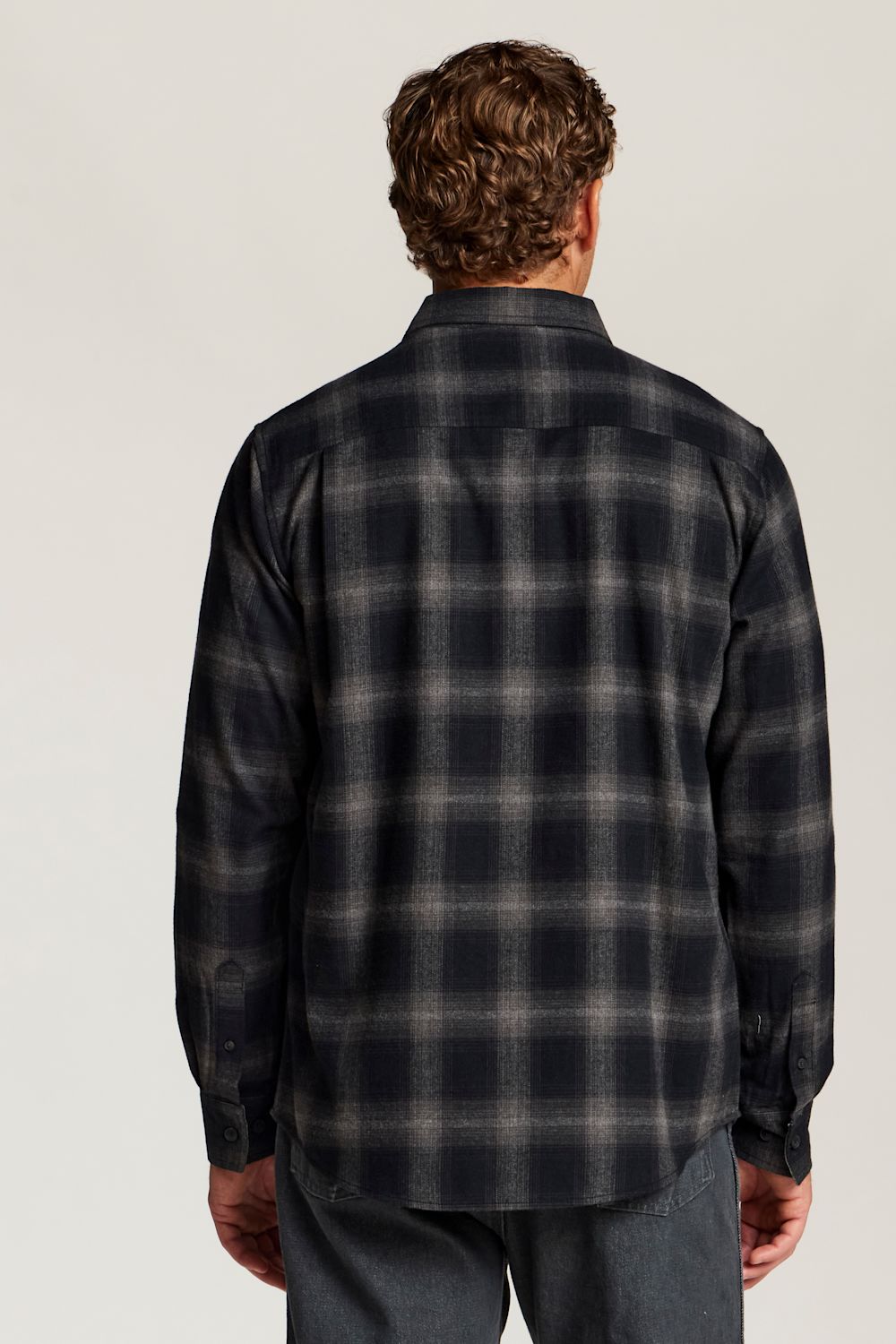 A man viewed from behind wearing a dark navy and gray plaid flannel shirt, paired with gray jeans, against a light gray studio background.