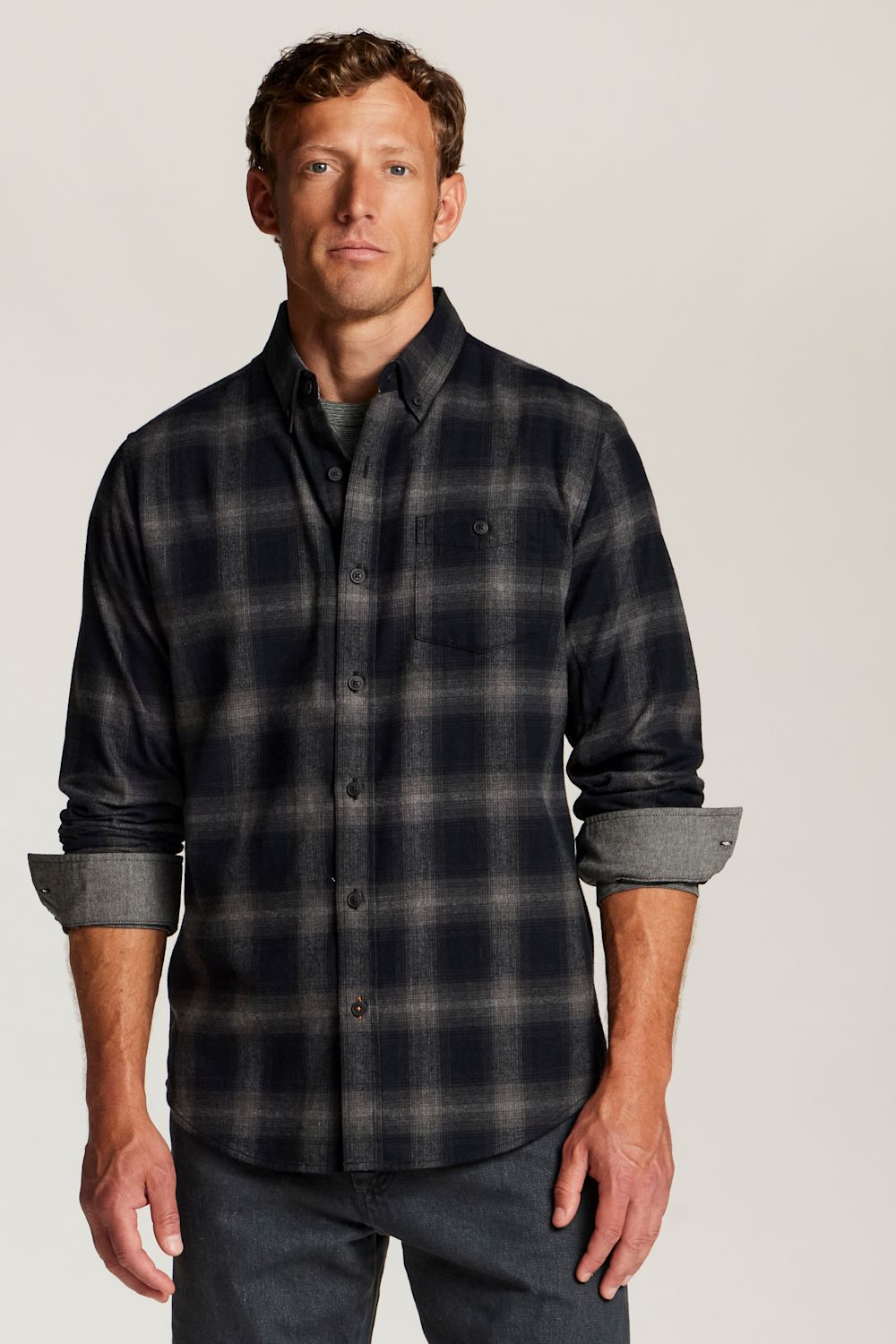 A man wearing a dark charcoal and gray plaid button-down shirt with rolled-up sleeves, paired with dark jeans, against a solid light gray background.