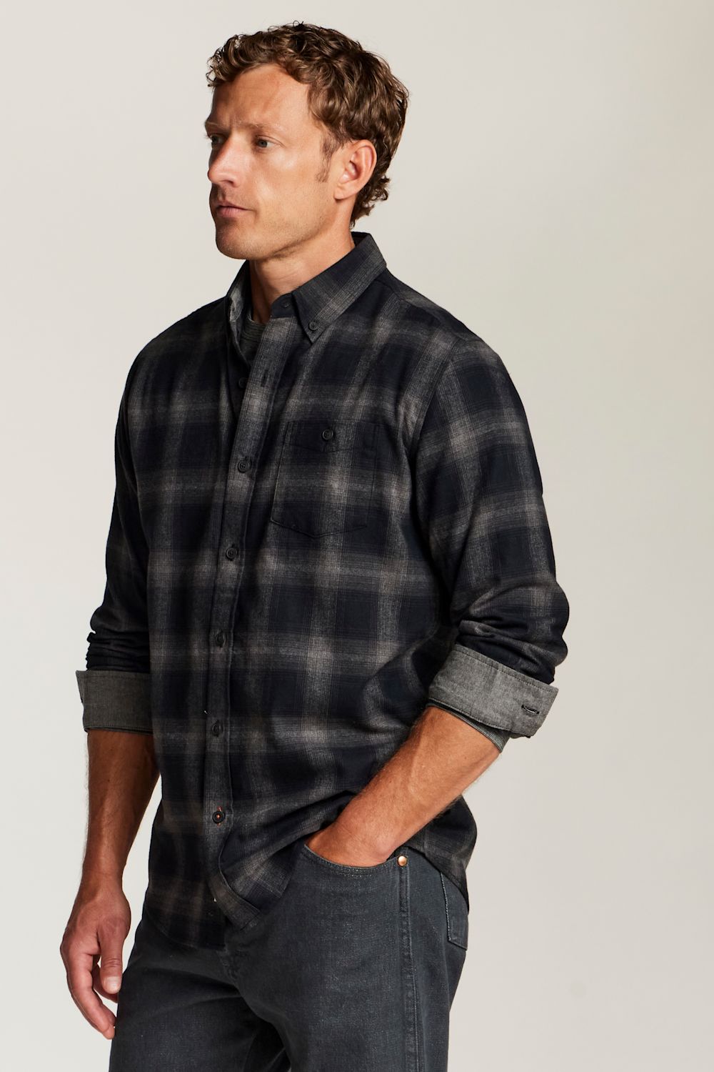 A man wearing a dark navy and gray plaid button-down shirt with rolled-up sleeves, paired with dark jeans, photographed against a neutral light gray studio background.
