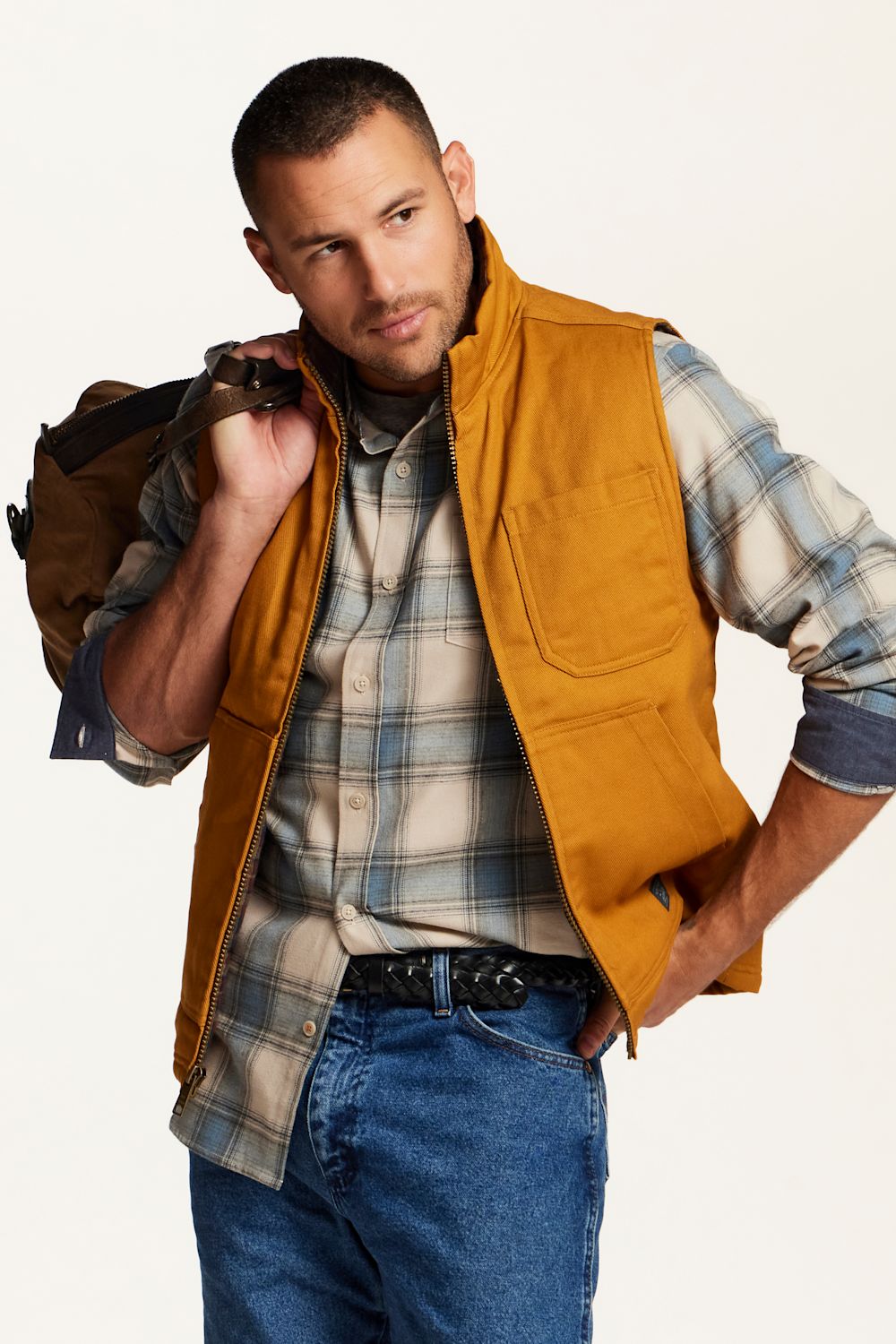 A man wearing a mustard yellow utility vest over a blue and beige plaid flannel shirt, paired with blue jeans and a brown backpack, against a plain off-white background.