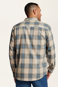 A man viewed from the back wearing a long-sleeve plaid button-up shirt in beige, blue, and gray, paired with blue jeans, against a plain off-white background.