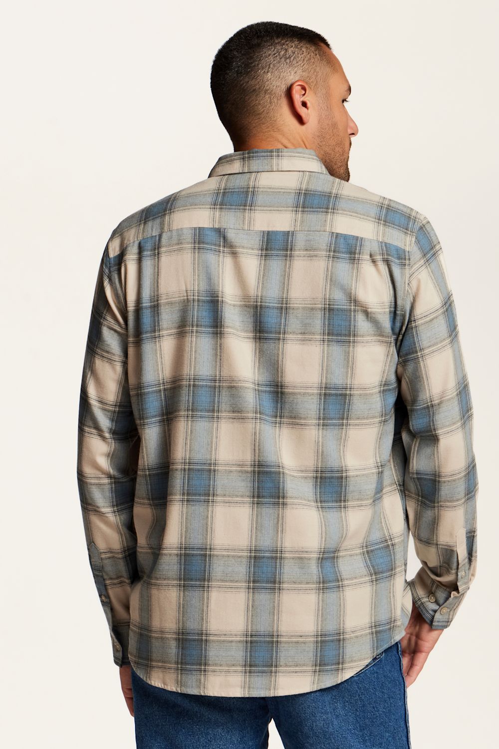 A man viewed from the back wearing a long-sleeve plaid button-up shirt in beige, blue, and gray, paired with blue jeans, against a plain off-white background.