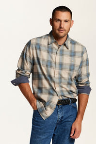 A man wearing a beige and blue plaid button-down shirt with rolled-up sleeves, tucked into blue jeans with a black belt, against a plain white studio background.