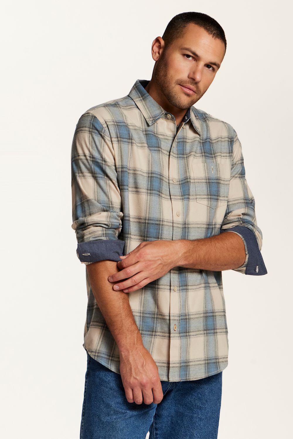 A man wearing a long-sleeve button-down plaid shirt in beige, blue, and gray, paired with blue jeans, photographed against a clean off-white studio background.