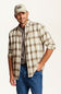A man wearing a beige and brown plaid button-down shirt over a dark gray t-shirt, blue jeans, and a beige baseball cap, posed against a solid off-white background.