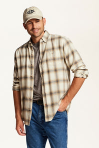A man wearing a beige and brown plaid button-down shirt over a dark gray t-shirt, blue jeans, and a beige baseball cap, posed against a solid off-white background.