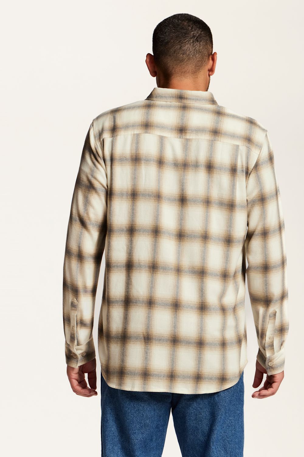 A man viewed from the back wearing a long-sleeve plaid button-up shirt in beige and brown with blue jeans, set against a plain white studio background.