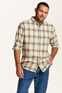 A man wearing a beige and brown plaid button-down shirt with rolled-up sleeves, paired with blue jeans, standing against a plain white studio background.