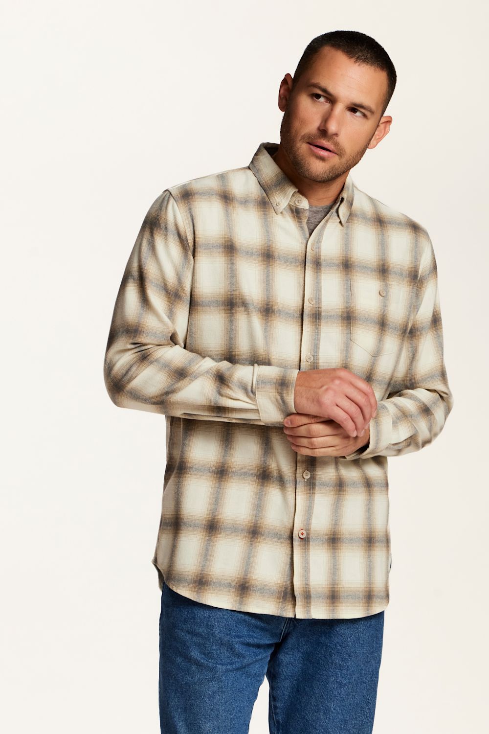 A man wearing a beige and brown plaid button-down shirt with blue jeans, standing against a plain white studio background.
