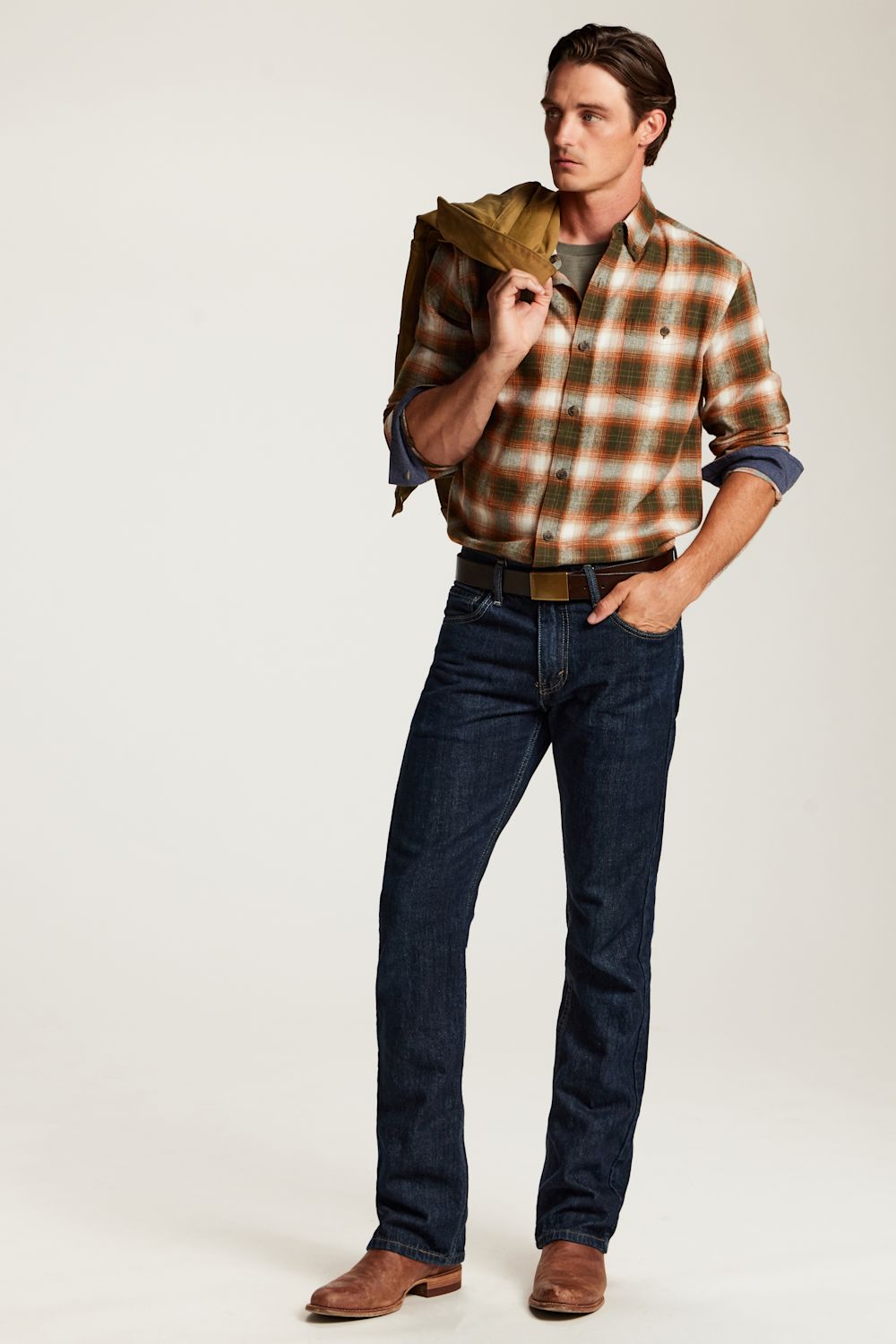 A man modeling a brown and orange plaid button-down shirt with rolled sleeves, dark blue jeans, and brown boots, while holding a brown jacket over his shoulder against a light gray studio background.
