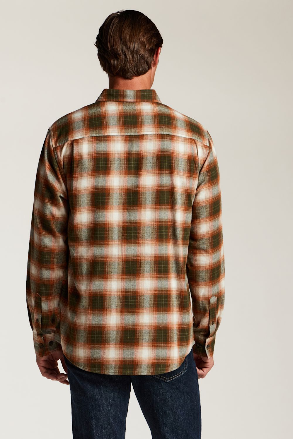 A man viewed from behind wearing a long-sleeve plaid button-up shirt in brown, orange, and cream tones, paired with dark blue jeans, against a plain light gray studio background.